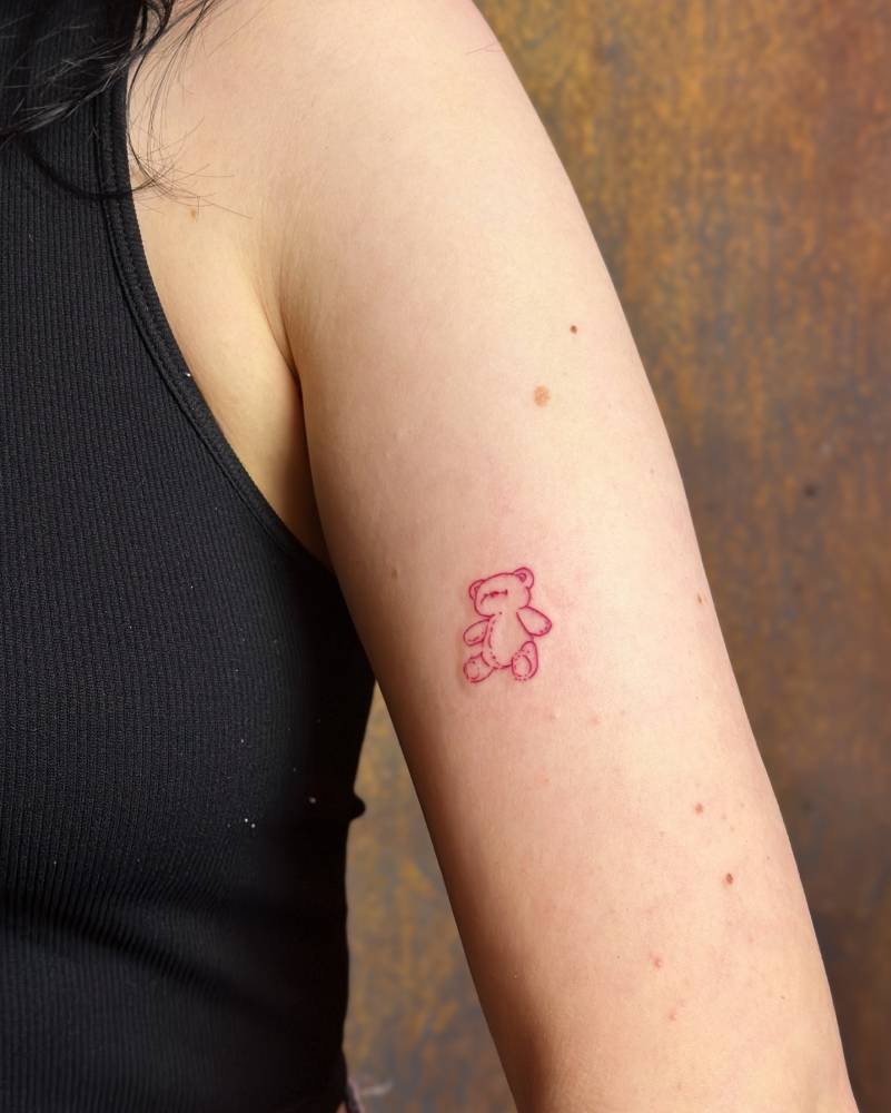 Fine line style red teddy bear tattoo placed on the upper arm.