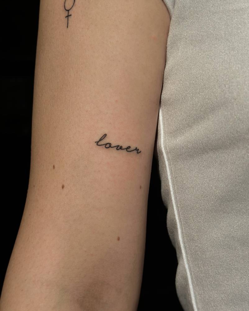 Handwritten lettering tattoo of the word "lover" located on the bicep.