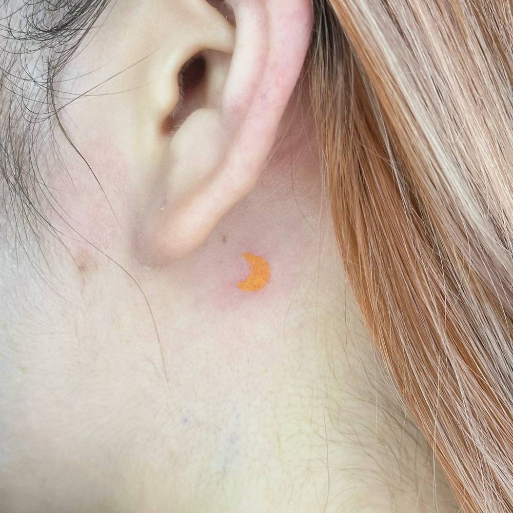 Tiny yellow crescent moon tattoo hand poked behind the ear.