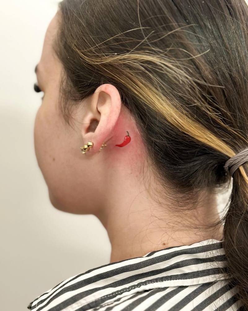 Tiny red chili tattoo placed behind the ear, illustrative style.