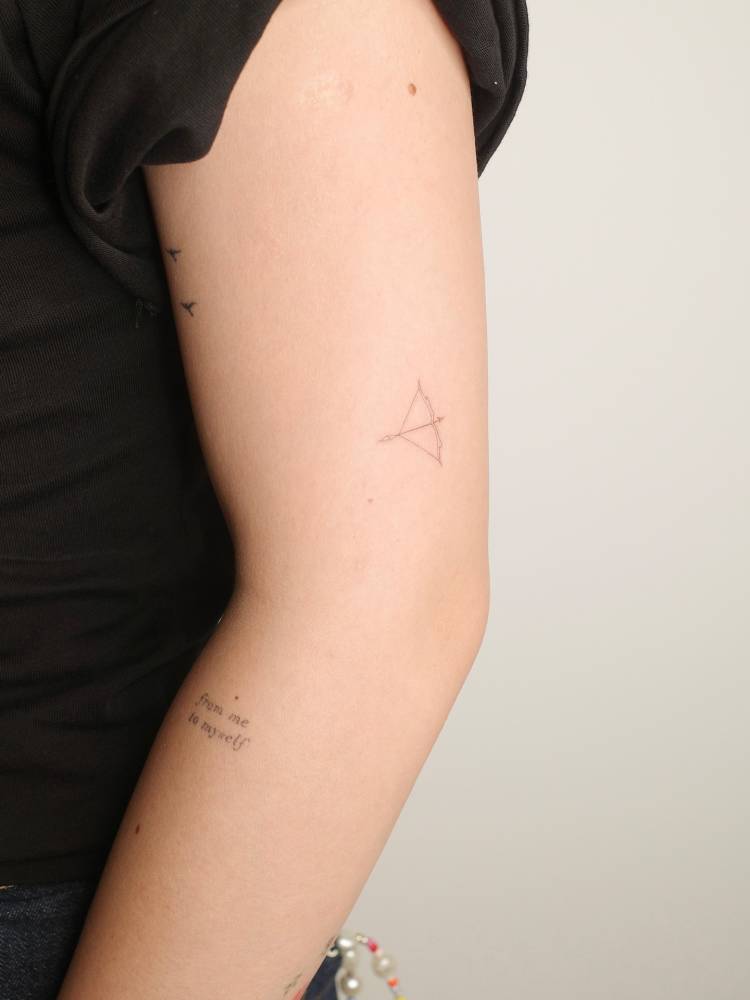 Bow and arrow tattoo located on the upper arm, fine line style.