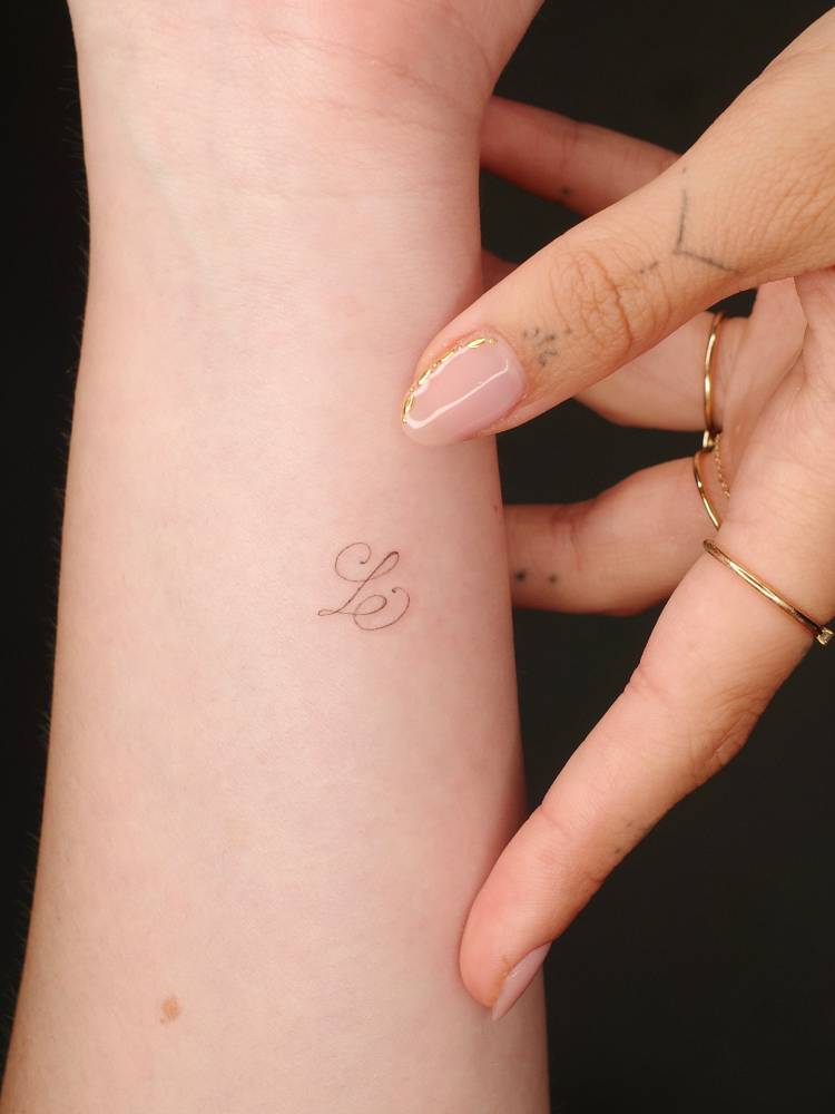 Tiny letter "L" in script font tattooed on the wrist.