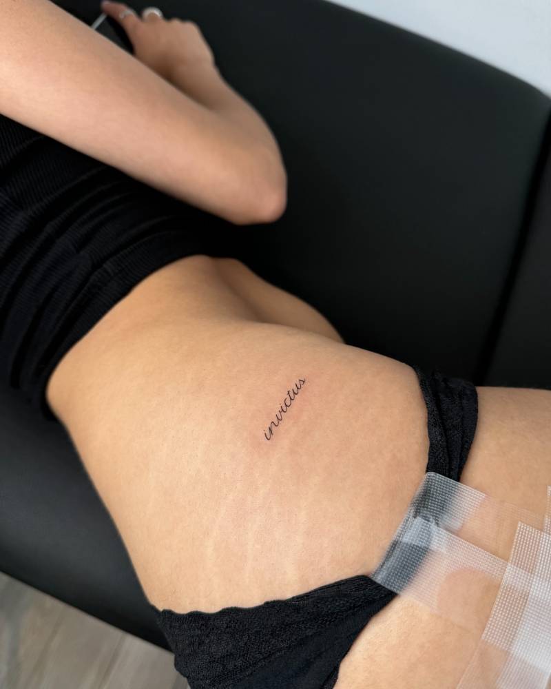 Handwritten font word "invictus" tattooed on the hip.