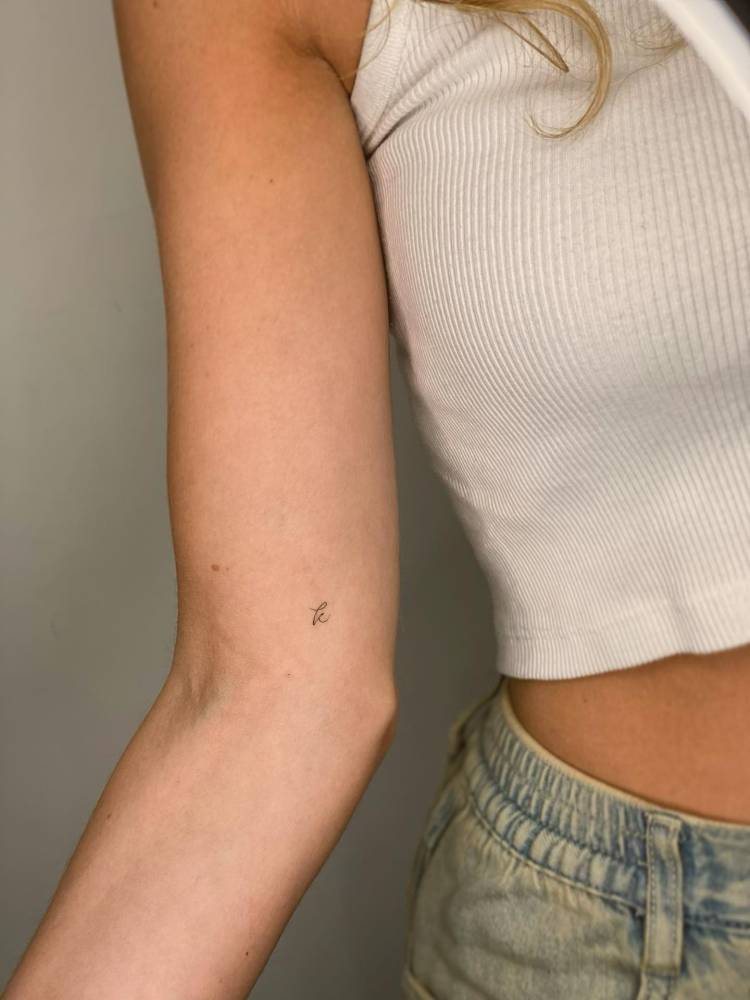 Tiny handwritten font letter "k" tattooed on the inner arm.
