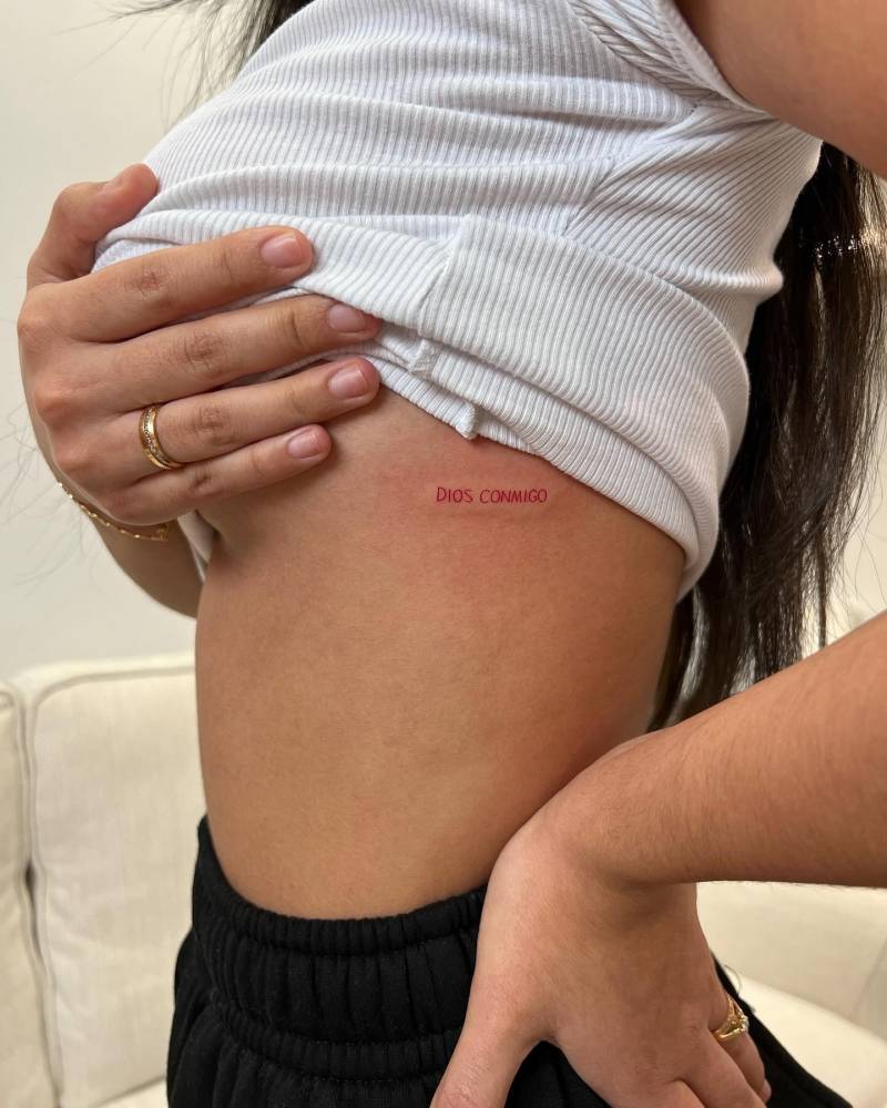 "Dios conmigo" lettering tattoo in red ink located on the rib.