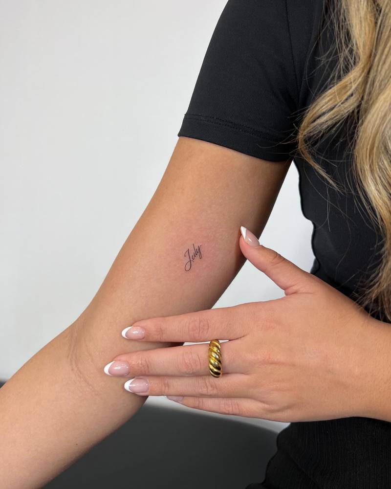 "July" lettering tattoo in script font located on the inner arm.