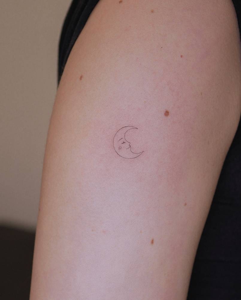 Crescent moon face tattoo located on the upper arm, fine line style.