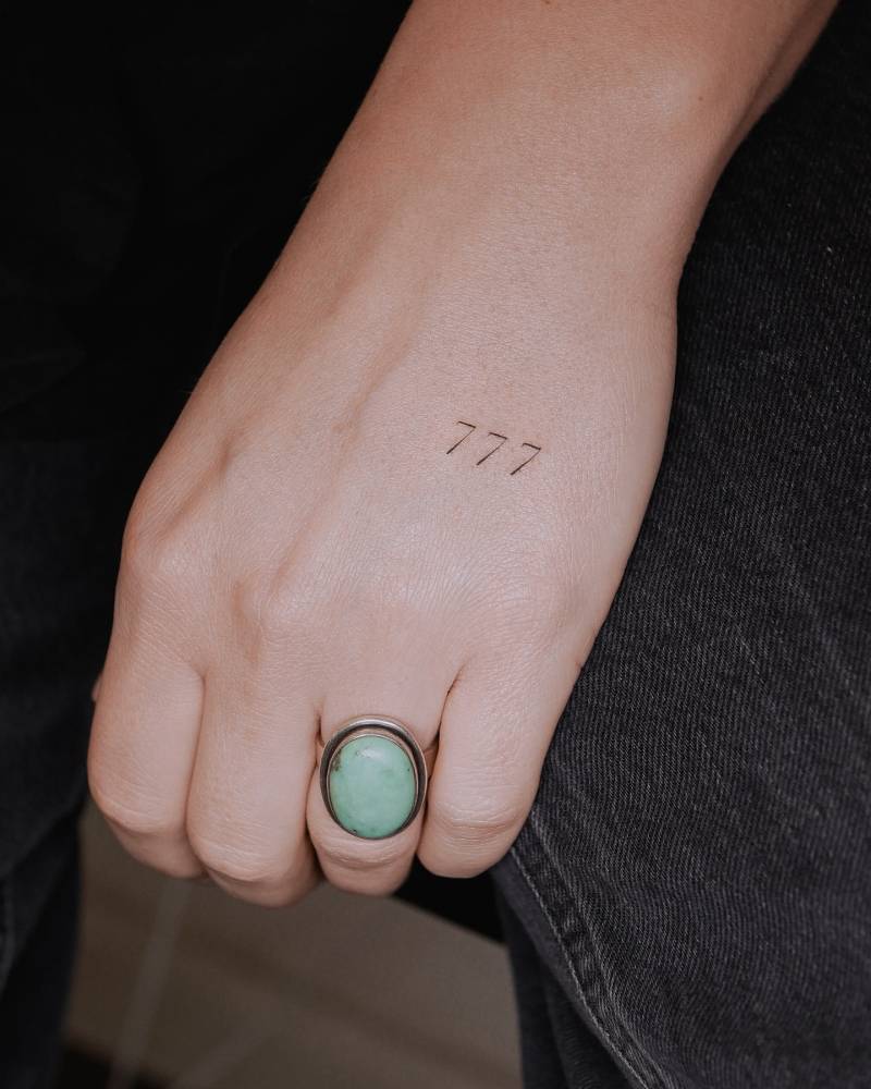 Angel number "777" tattoo in serif font located on the hand.