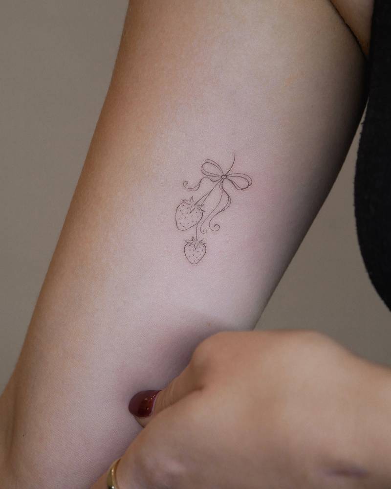Strawberries and bow tattoo in fine line style located on the inner arm.