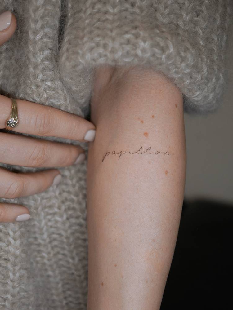 Handwritten lettering tattoo that says "papillon" located on the forearm.