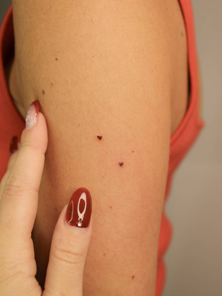 Tiny heart freckles tattoo located on the upper arm.