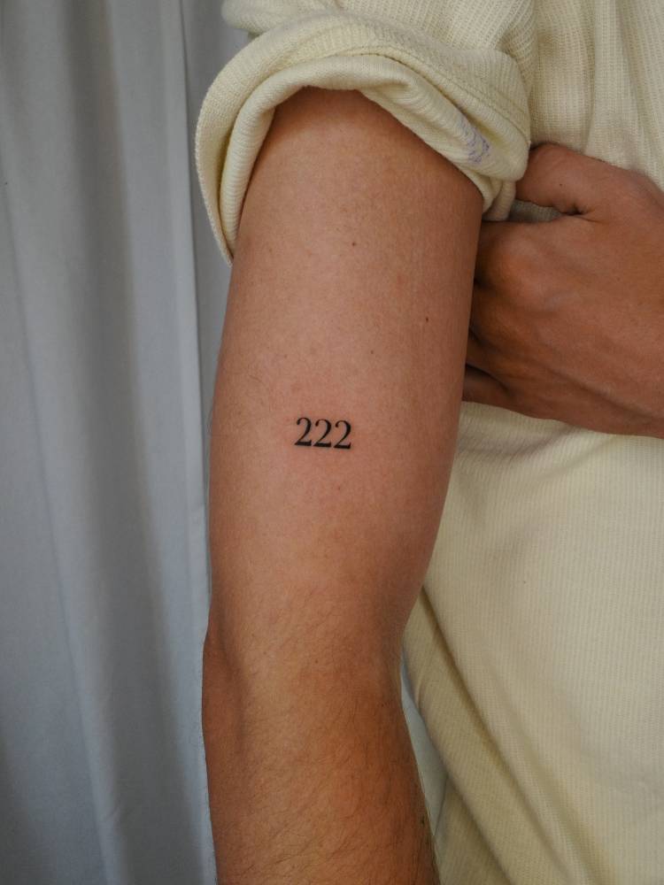 Angel number "222" in serif font tattooed on the upper arm.