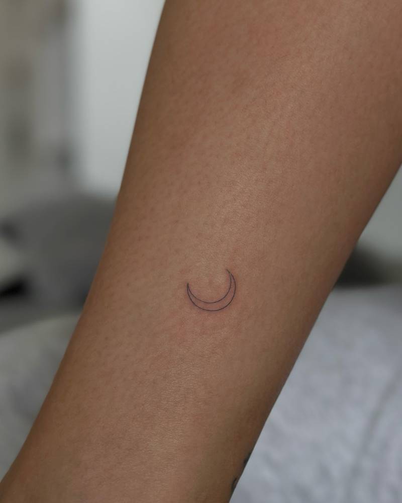 Tiny fine line crescent moon tattoo in blue ink located on the shin.