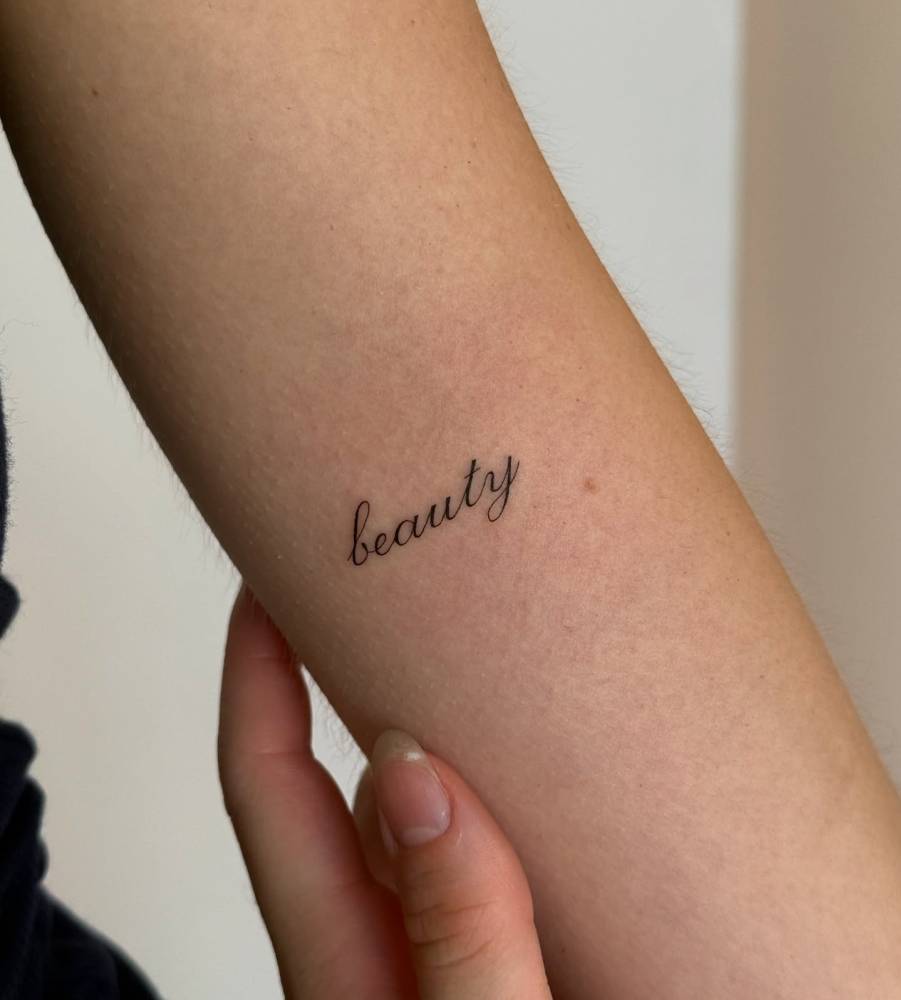 Word "beauty" lettering tattoo in script font located on the inner arm.