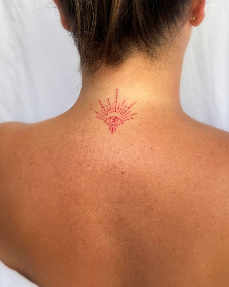 Ornamental evil eye tattoo in red ink located on the back of the neck.