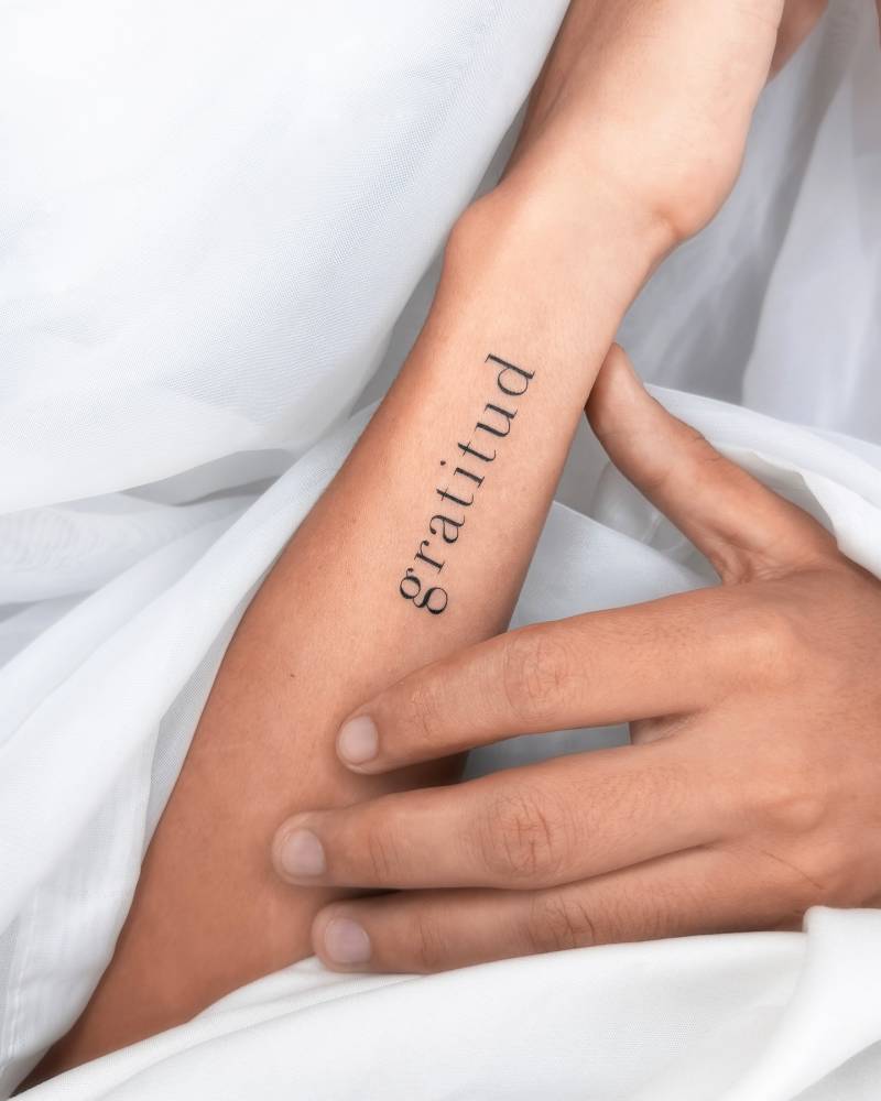 Serif font word "gratitud" lettering tattoo located on the wrist.