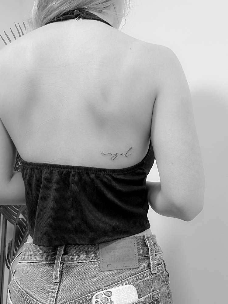 Handwritten lettering tattoo of the word "angel" placed on the back.