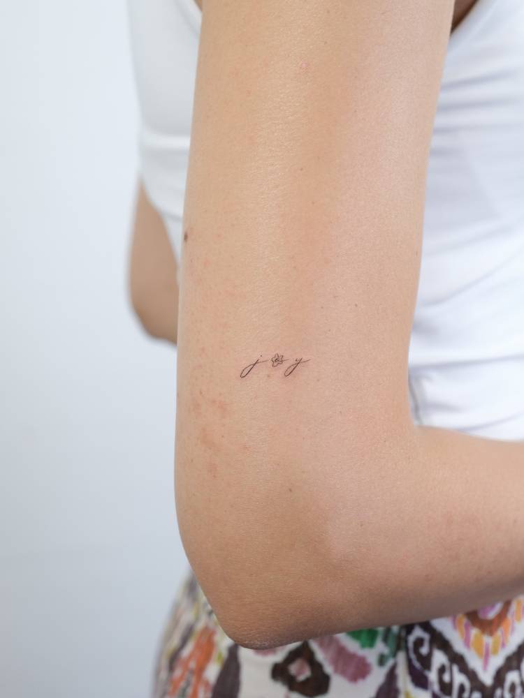 Word "joy" in handwritten font tattooed on the upper arm.