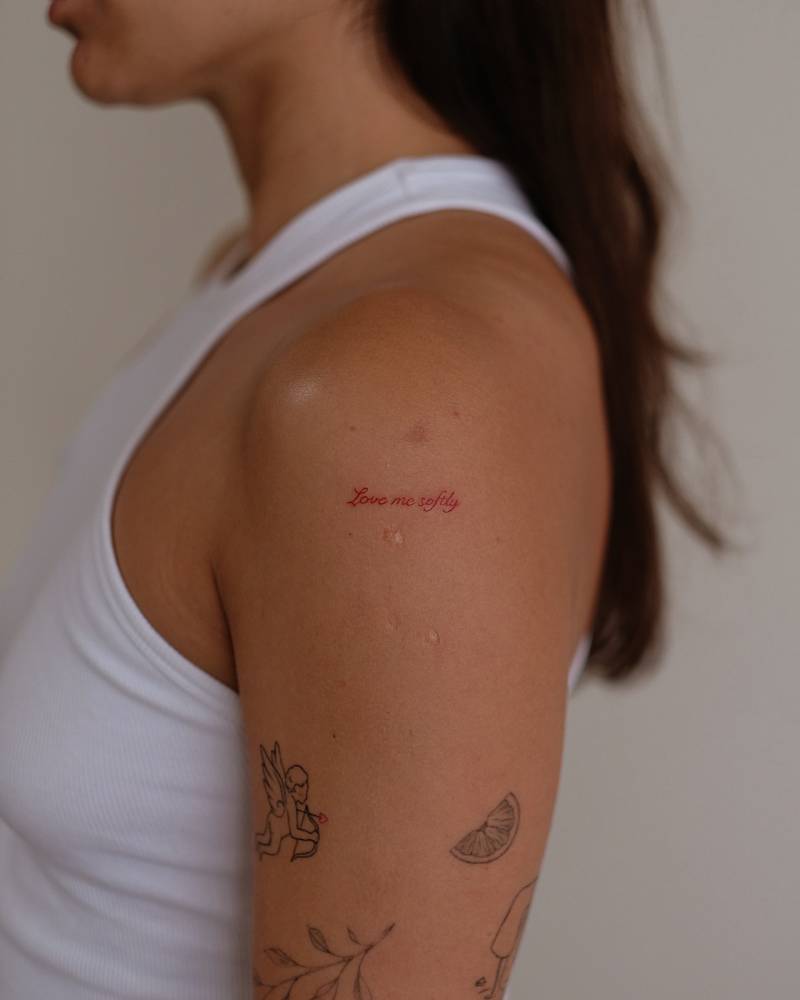 "Love me softly" lettering tattoo in red ink located on the upper arm.
