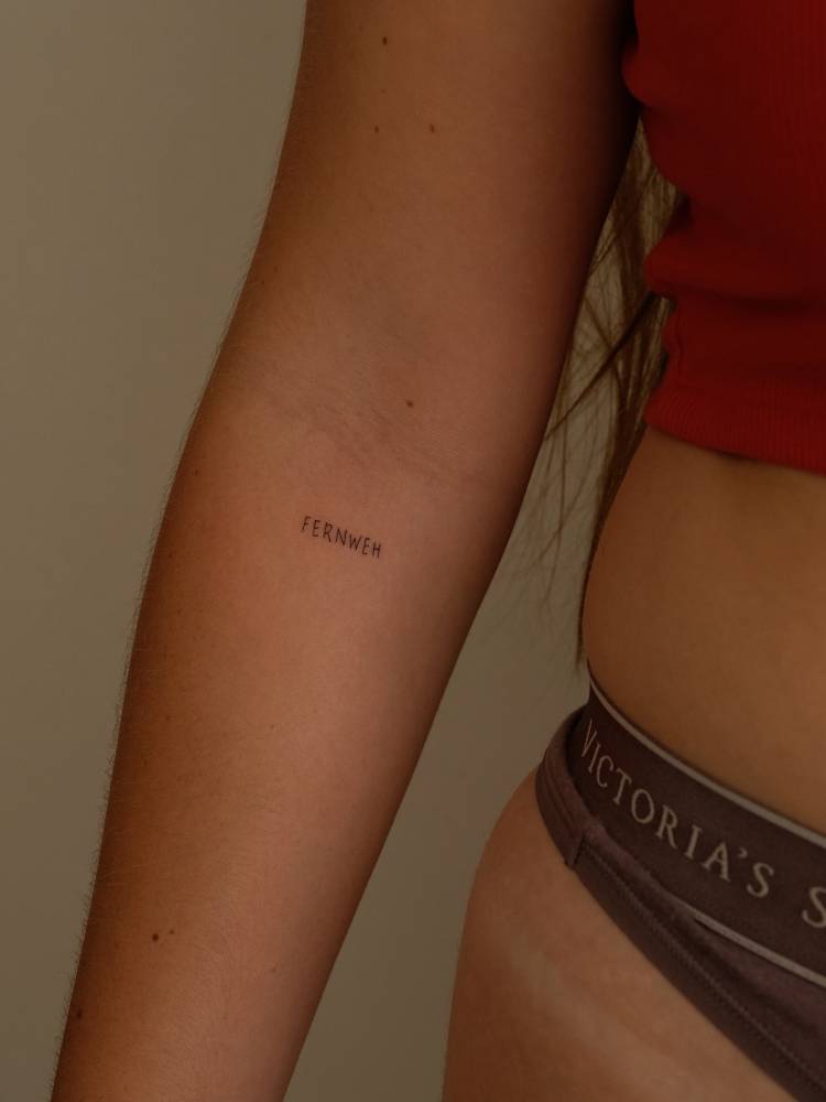 Word "fernweh" in handwritten font tattooed on the inner forearm.