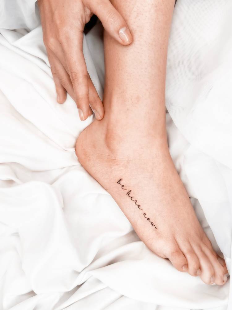 Lettering tattoo that says "be here now" located on the foot.