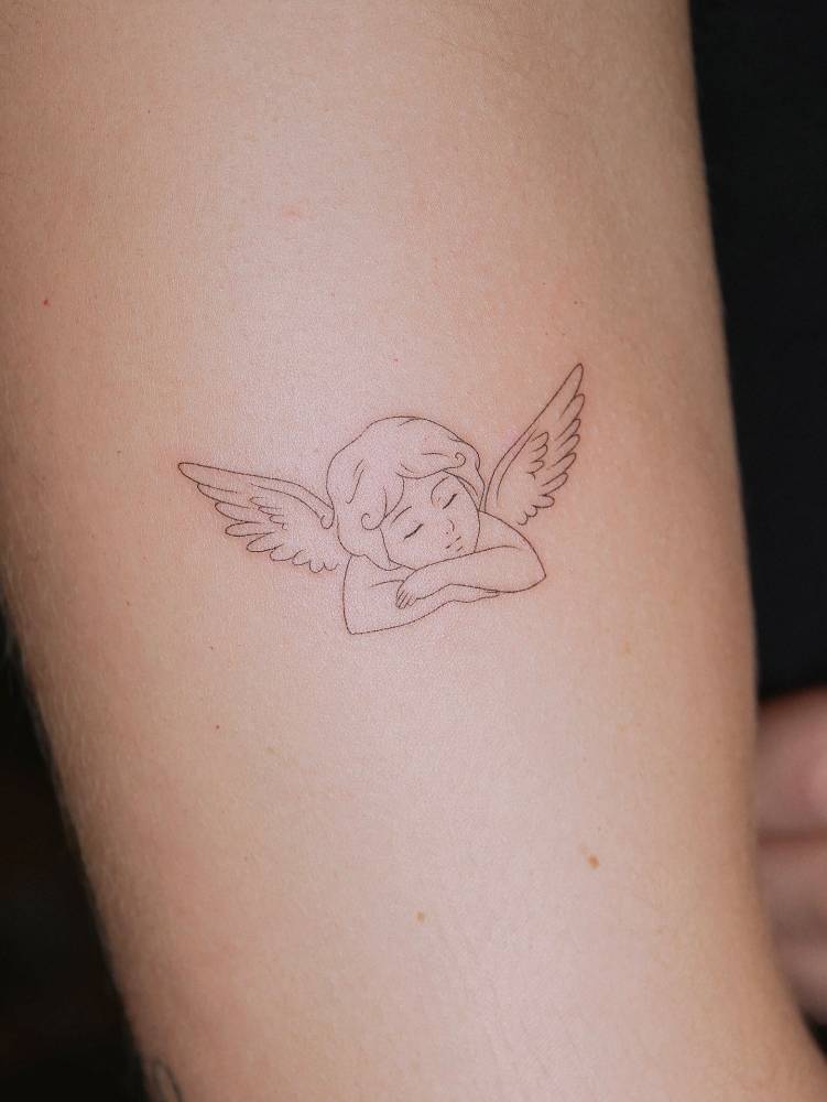Sleeping cherub tattoo in fine line style located on the upper arm.