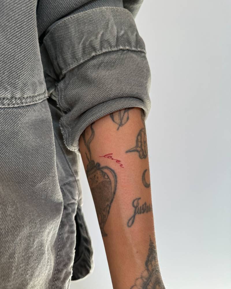Lettering tattoo of the word "lover" in red ink located on the forearm.