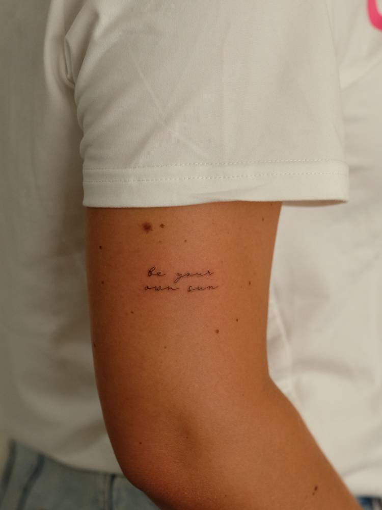 Handwritten font "be your own sun" lettering tattoo located on the upper arm.