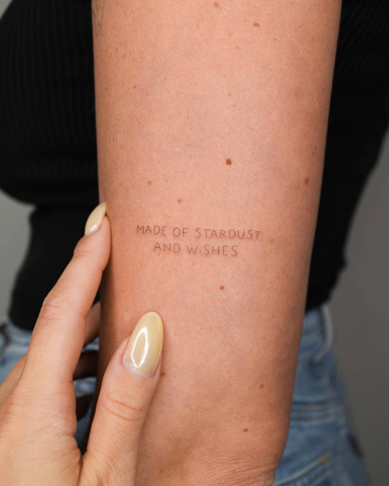 "Made of stardust and wishes" lettering tattoo handwritten on the upper arm.