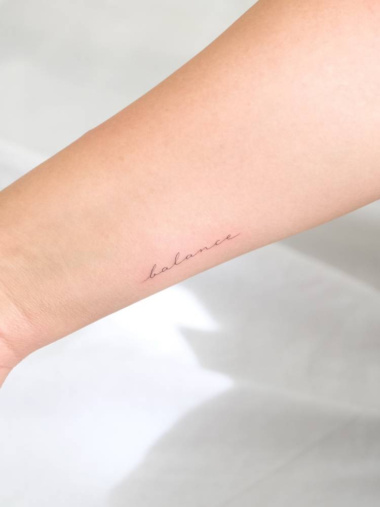 Handwritten font word "balance" tattooed on the wrist.