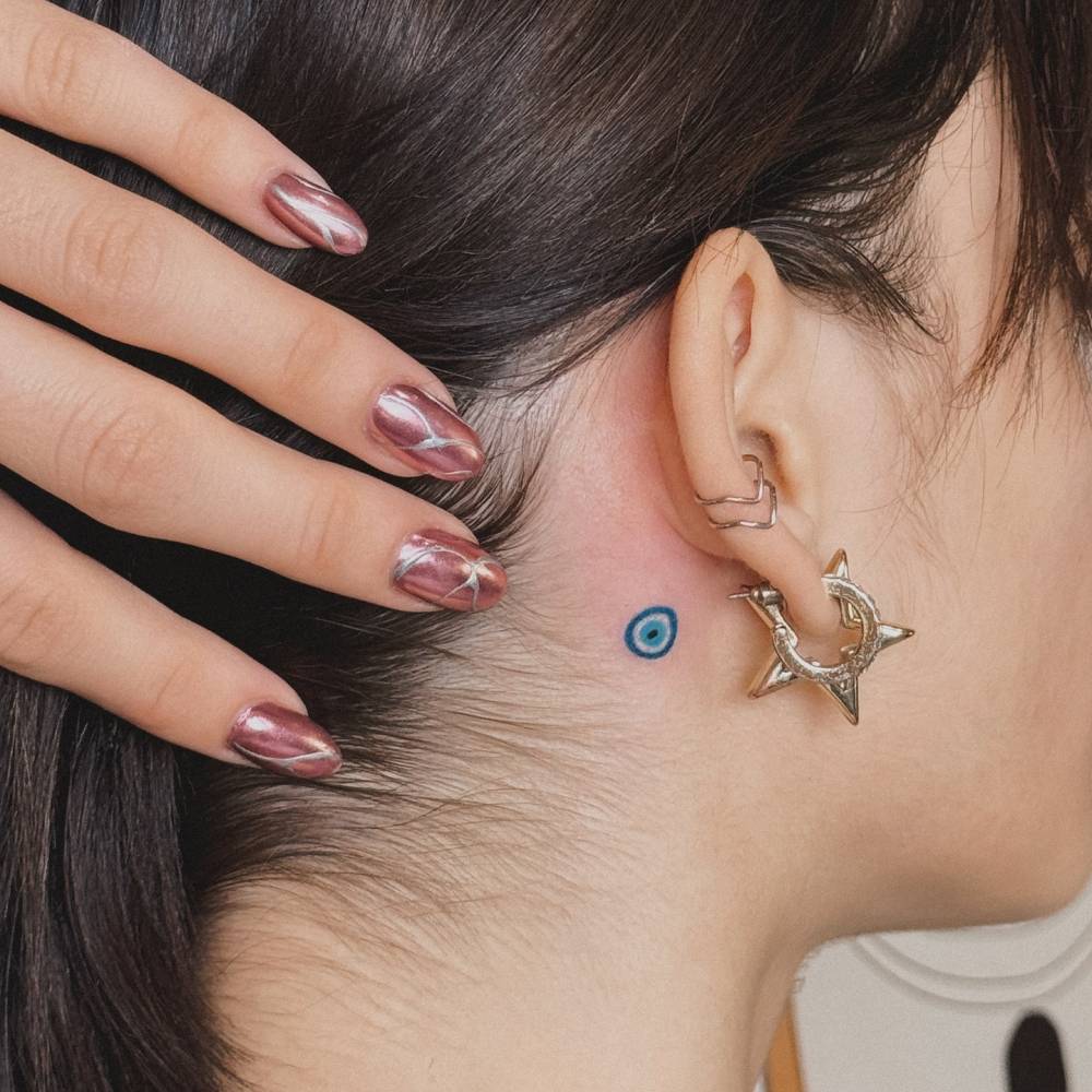 Tiny minimalistic Nazar tattoo located behind the ear.