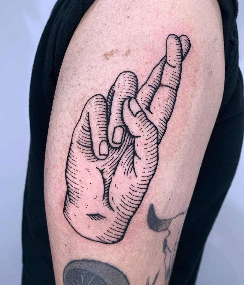 Crossed fingers tattooed on the upper arm in engraving style.