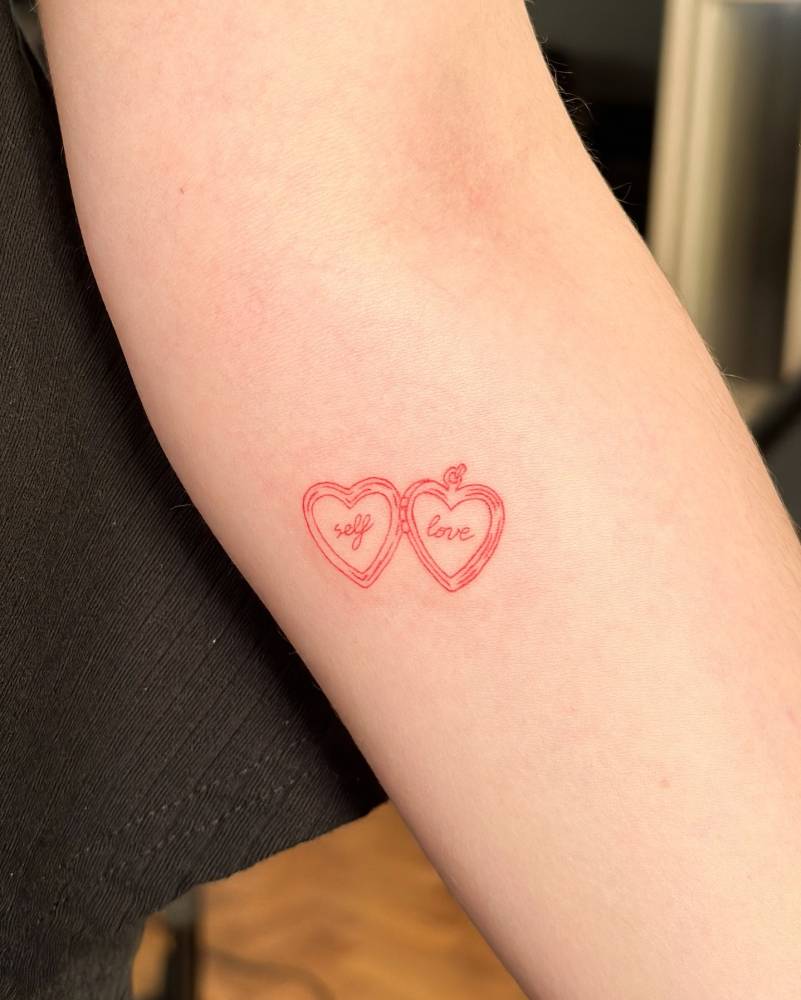 "Self love" heart pendant tattoo in red ink located on the inner forearm.
