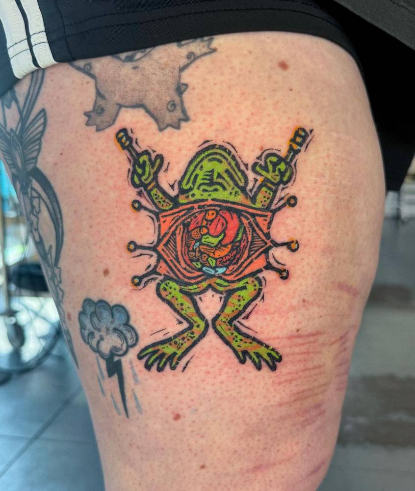 Cartoon style open frog tattoo located on the thigh.