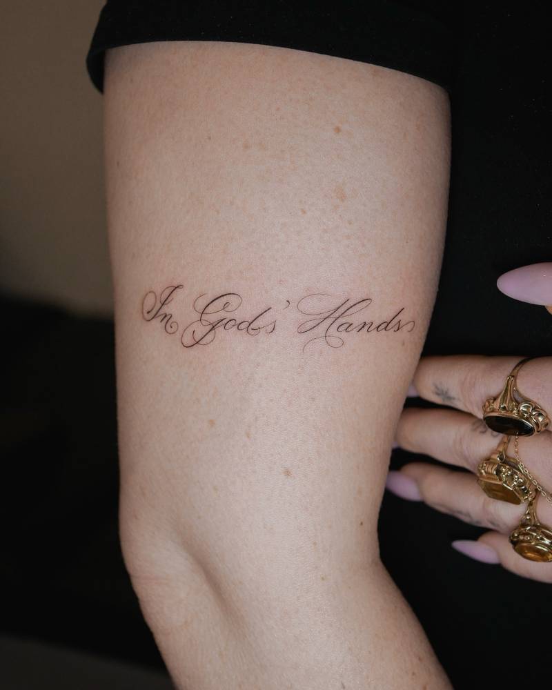"In God's hands" lettering tattoo located on the upper arm.