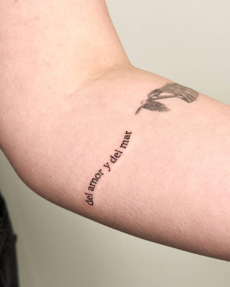 "Del amor y del mar" lettering tattoo located on the inner forearm.