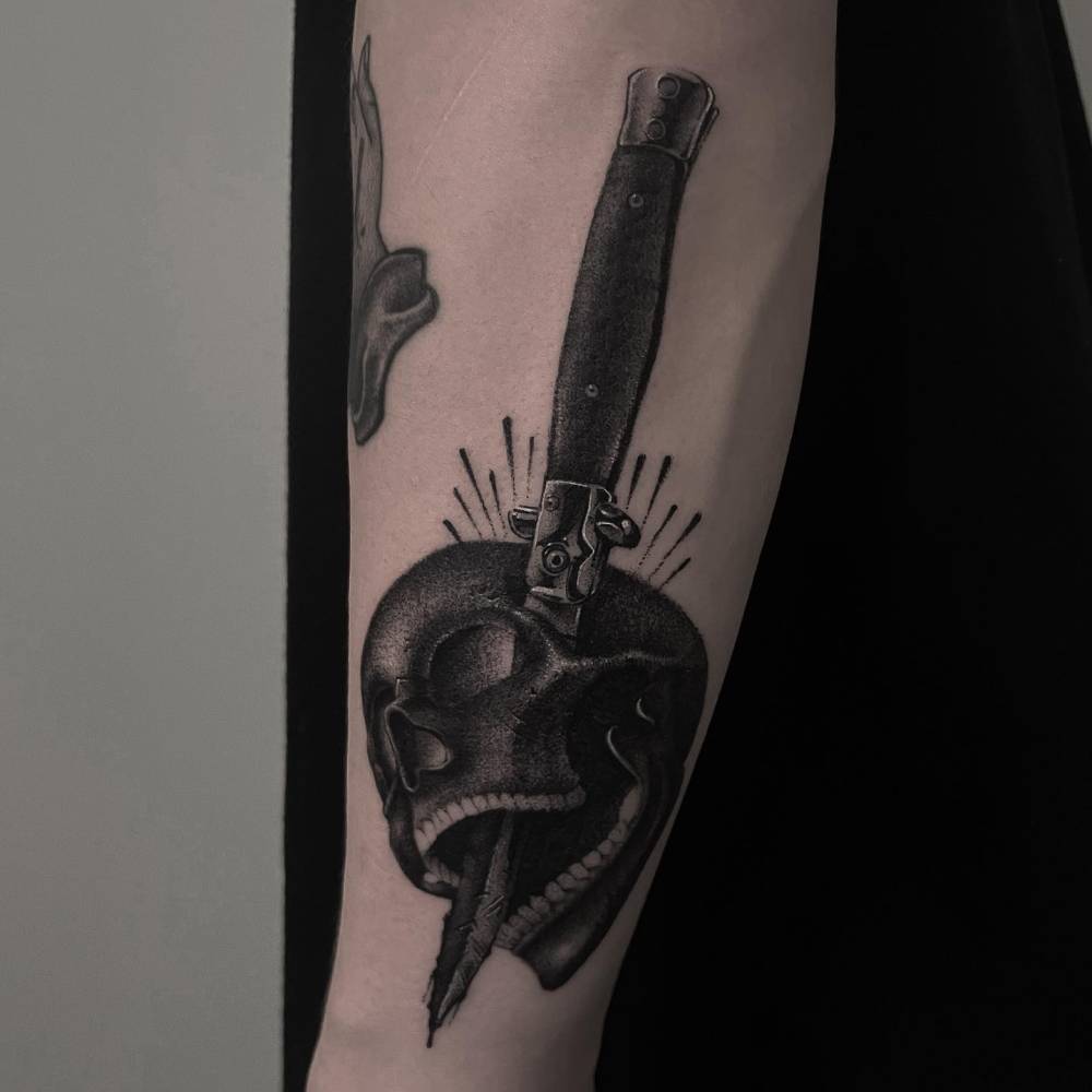 Black and grey style skull and knife tattoo located on the forearm.