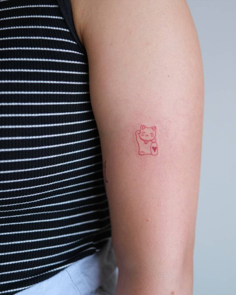 Fine line style Maneki-neko tattoo in red ink located on the upper arm.