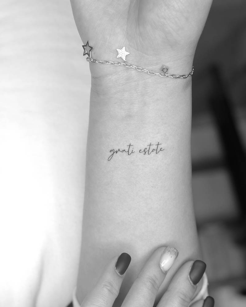"Grati estote" lettering tattoo in handwritten font located on the wrist.