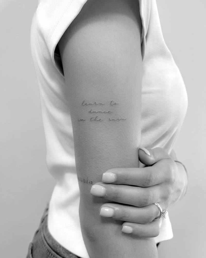 "Learn to dance in the rain" lettering tattoo located on the upper arm.