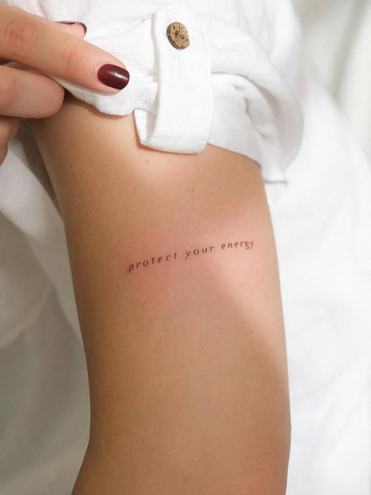 "Protect your energy" lettering tattoo located on the upper arm.