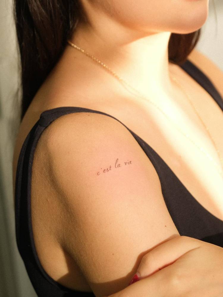 "C'est la vie" lettering tattoo located on the shoulder.