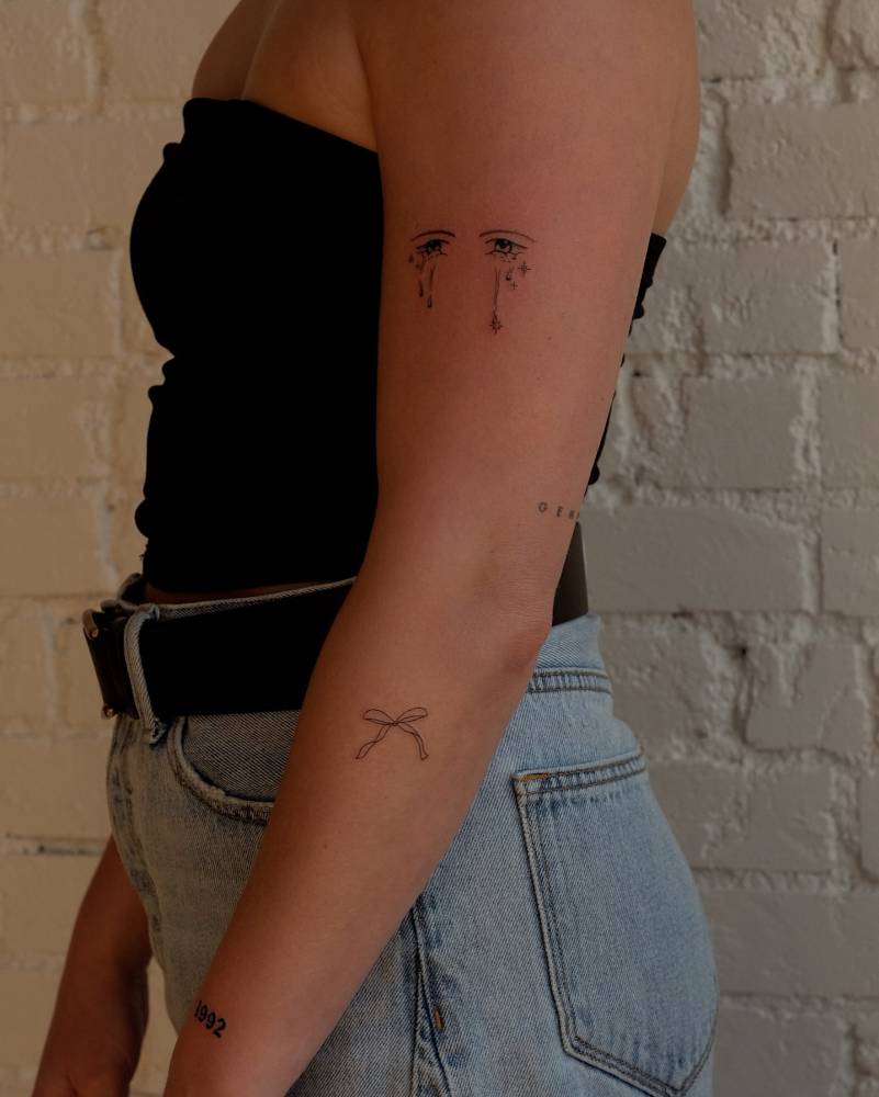 Crying eyes and bow tattoos located on the arm.