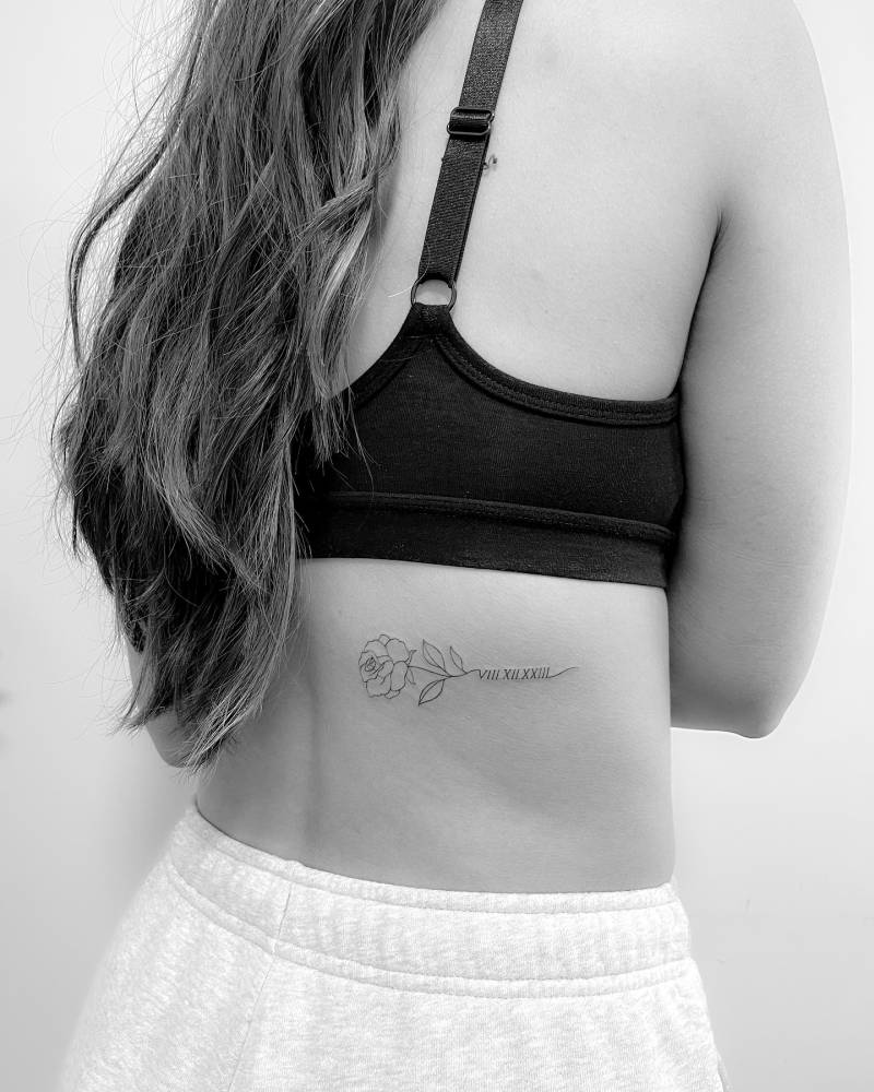 Fine line style rose and date in roman numerals tattoo located on the back.