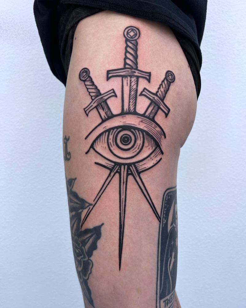 Swords and eye tattoo in engraving style located on the thigh.