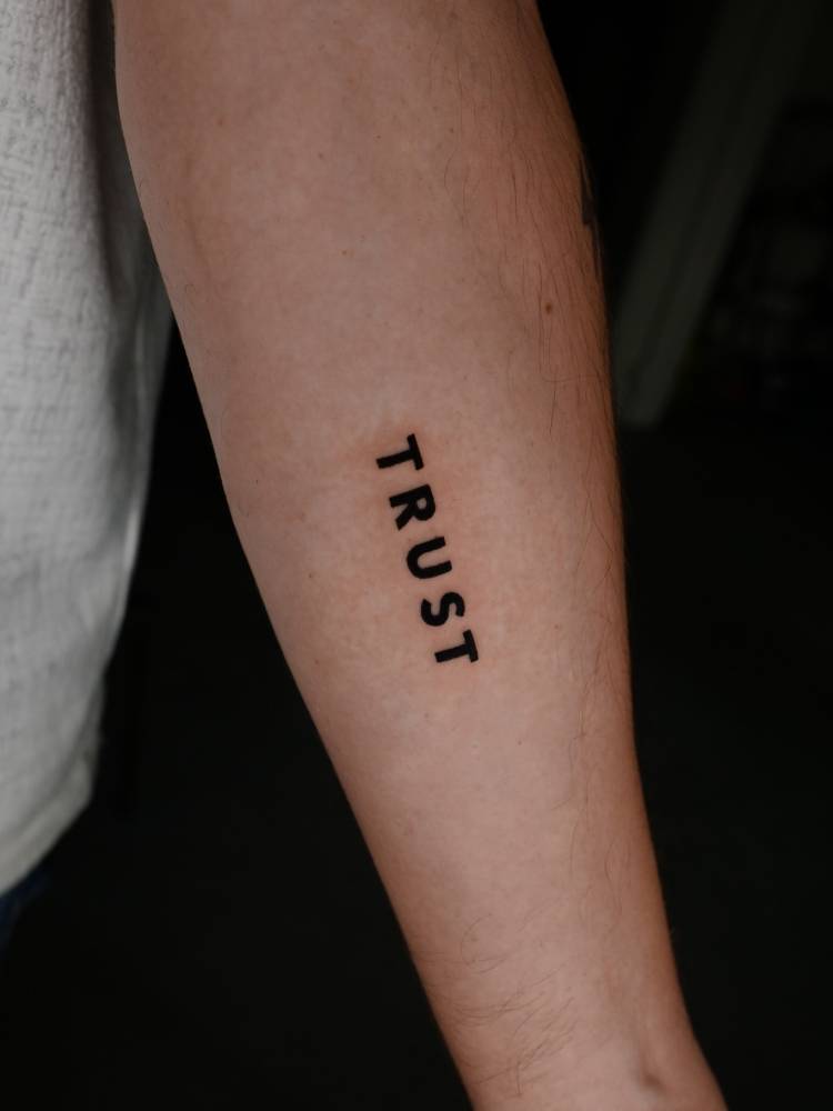Lettering tattoo of the word "trust" located on the inner forearm.