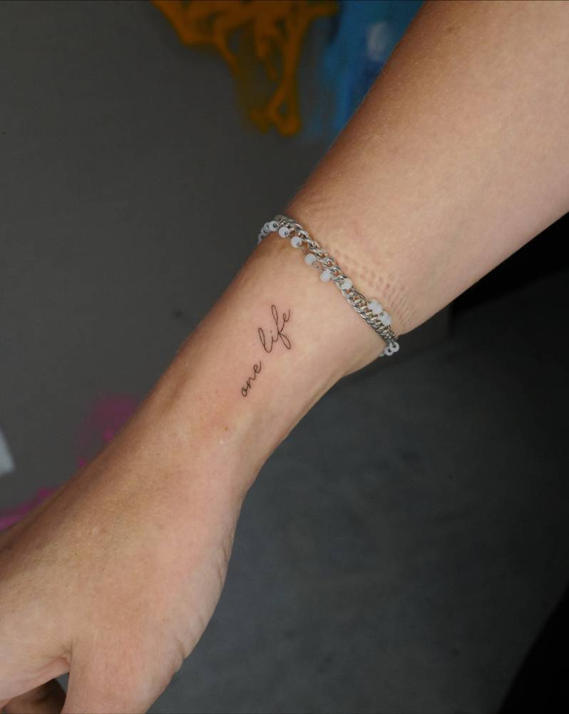 Handwritten font "one life" lettering tattoo located on the wrist.
