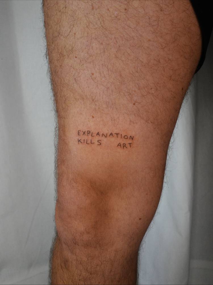 "Explanation kills art" lettering tattoo located on the thigh.