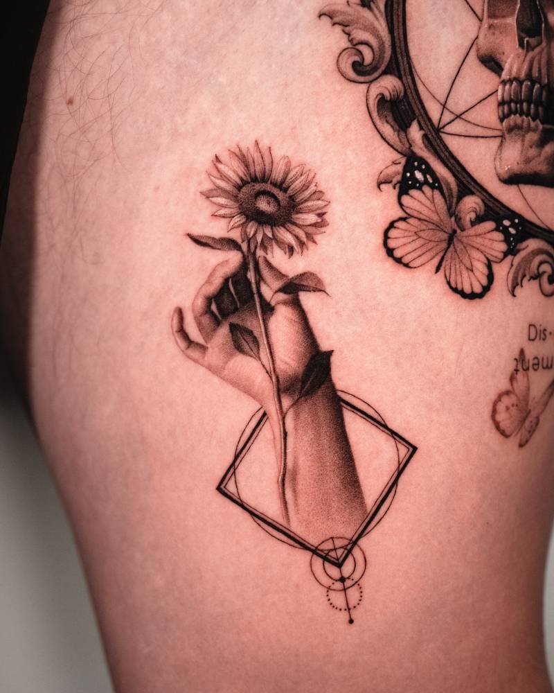 Hand holding a sunflower tattooed on the thigh, micro-realistic style.