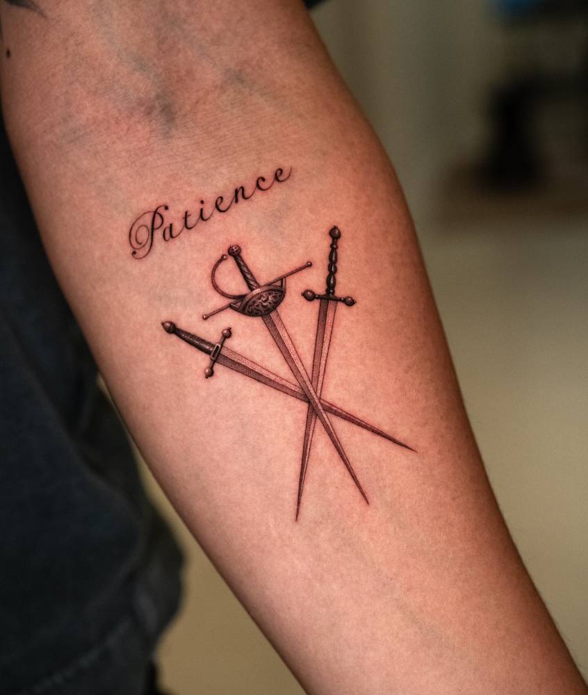 Word "patience" and three swords tattooed on the inner forearm.